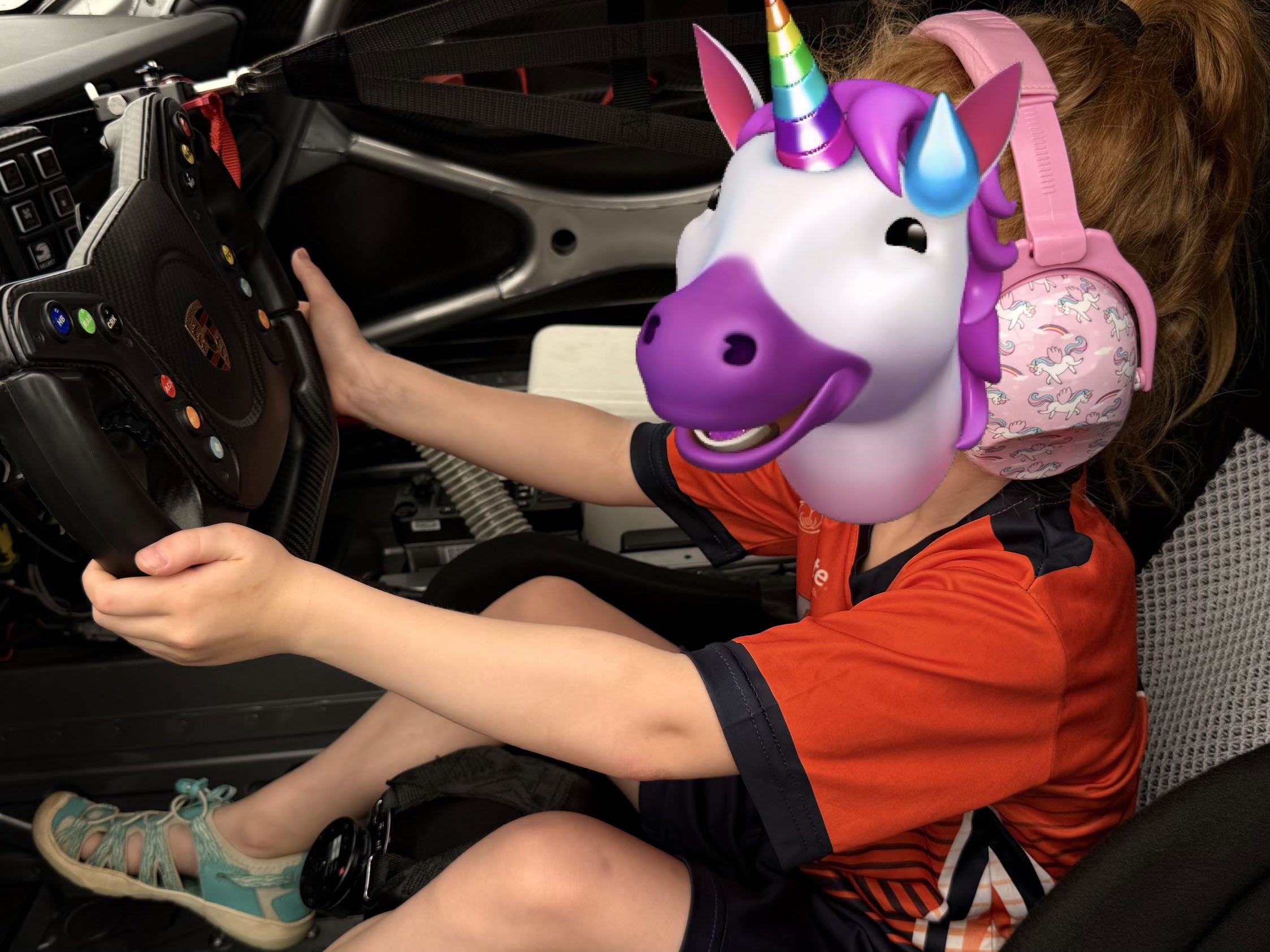 A child in a soccer uniform with her face censored by a unicorn emoji sitting in the driver’s seat of a Porsche Sprint Cup car holding the steering wheel